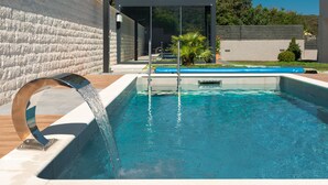 A modern outdoor swimming pool with a water feature, surrounded by a stone wall and a wooden deck. - WISH Apartments (Korcula)