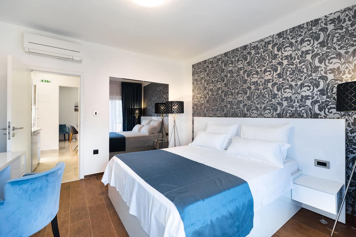 Ein Hotelzimmer mit einem großen Bett, einem Schreibtisch, einem Stuhl und einem Spiegel. Eine Tür führt zu einem Badezimmer und einem Flur.