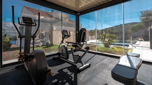 A gym with exercise equipment and a view of a residential area through large windows. - WISH Apartments (Korcula)