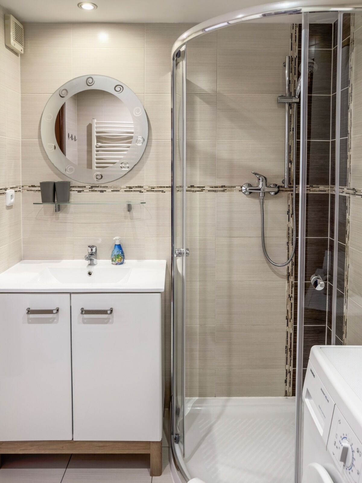 Un baño moderno con ducha de vidrio, un espejo redondo, un lavamanos blanco y una lavadora integrada.