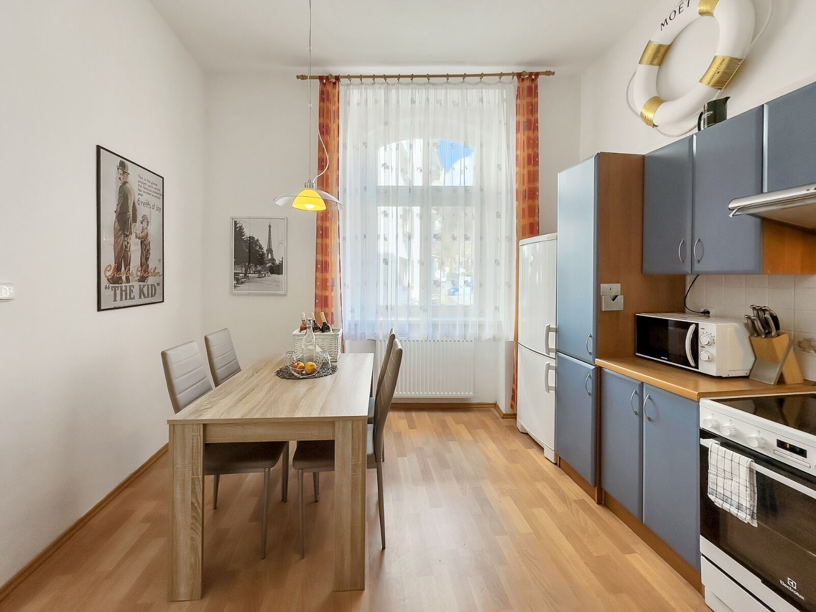 Une cuisine compacte équipée d’un four à micro-ondes, d’un four et de placards. Une table à manger avec des chaises est placée devant une fenêtre ornée de rideaux légers.