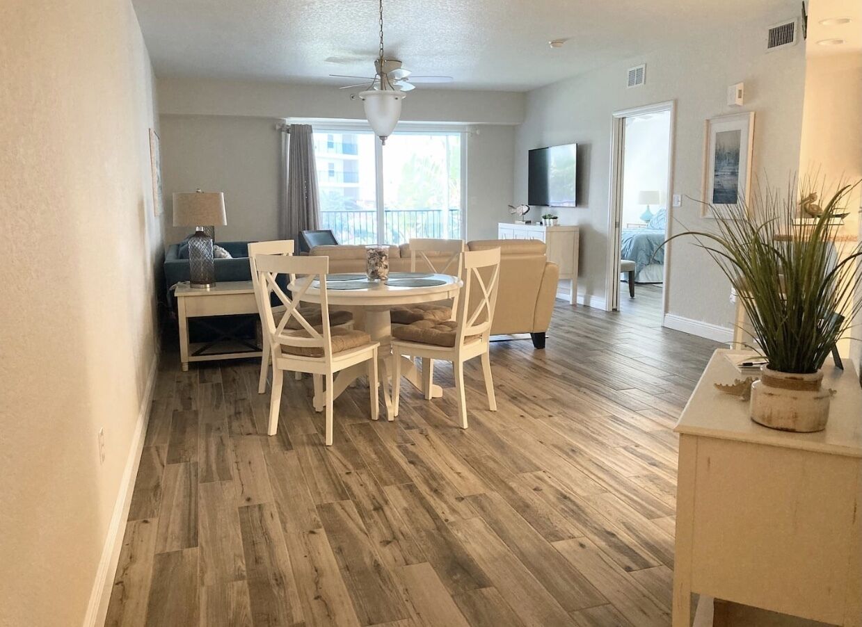 A spacious living room with a wooden floor, a dining area with white furniture, and a balcony with a view.
