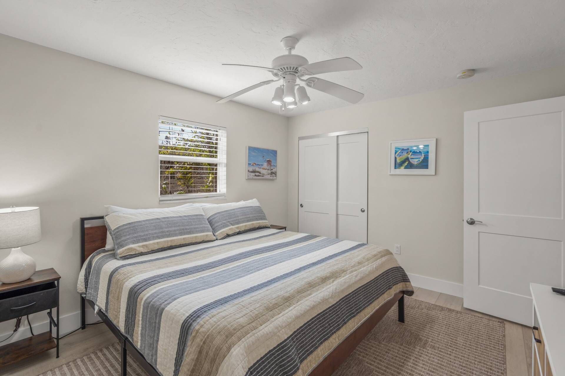 A bedroom with a bed, a ceiling fan, a window with blinds, a door, and a framed picture on the wall.