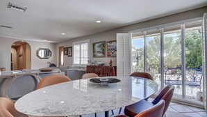 A spacious dining area with a marble table and modern chairs, large windows with a view of greenery, and a kitchen area with a wooden cabinet and artwork on the walls. - Canopy Cottage on Siesta Key (Siesta Key)