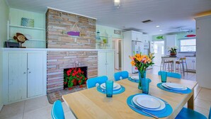 Un coin repas avec une table en bois dressée pour six personnes, des chaises bleues et une cheminée décorée de fleurs rouges.
