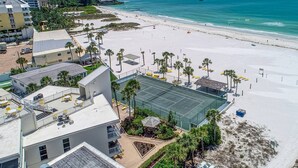 Vista aérea de un hotel frente a la playa, con cancha de tenis, palmeras y una playa de arena.
