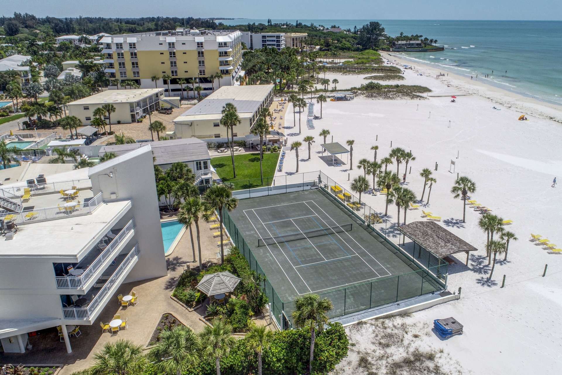 Un hotel frente a la playa con varias edificaciones, una cancha de tenis y una zona de piscina.