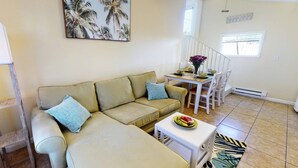 A living room with a beige sofa, a dining table with chairs, a small white table with a plate of fruit, and a tropical-themed painting on the wall.