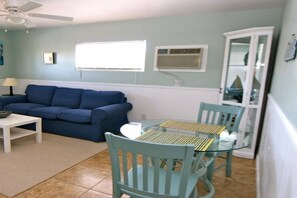Ein Wohnzimmer mit blauem Sofa, einem weißen Couchtisch und einem Esstisch mit Glasplatte und zwei Stühlen. Es gibt einen Deckenventilator, ein Fenster mit Jalousien und eine Klimaanlage an der Wand.