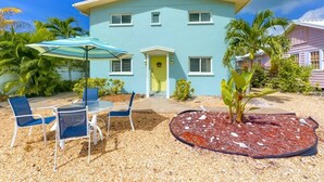Ein zweistöckiges Haus mit hellblauer Fassade, einer gelben Haustür und einem kleinen Veranda-Bereich. Vor dem Haus befindet sich eine Terrasse mit Tisch und Stühlen sowie ein kleiner Blumenkasten mit Mulch und Pflanzen.
