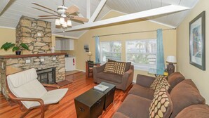 A spacious living room with a stone fireplace, a ceiling fan, and a large window with blinds.