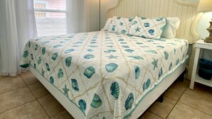 A bed with a quilted cover featuring a leaf pattern, a bedside table with a lamp, and a window with blinds.
