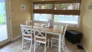 A dining area with a wooden table and white chairs, a small black table, and a window with blinds.