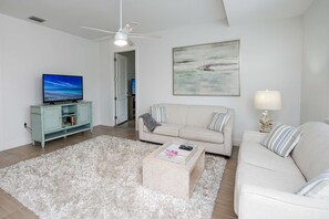 A living room with a beige sofa, a white rug, a coffee table, a TV on a light blue cabinet, and a ceiling fan.