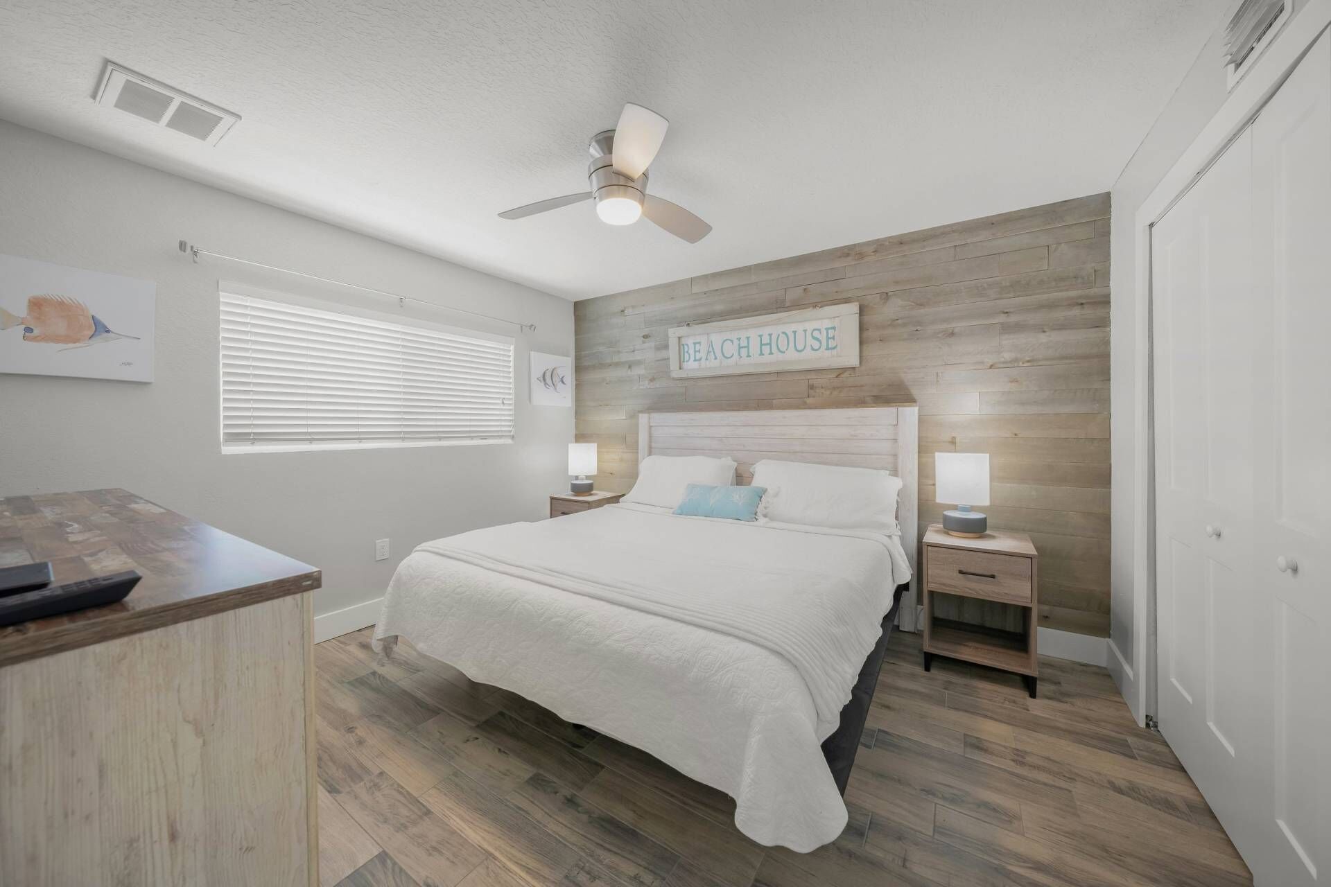 Quarto com cabeceira de madeira, cama com roupa de cama branca, duas mesas de cabeceira com luminárias, um ventilador de teto e uma janela com persianas.