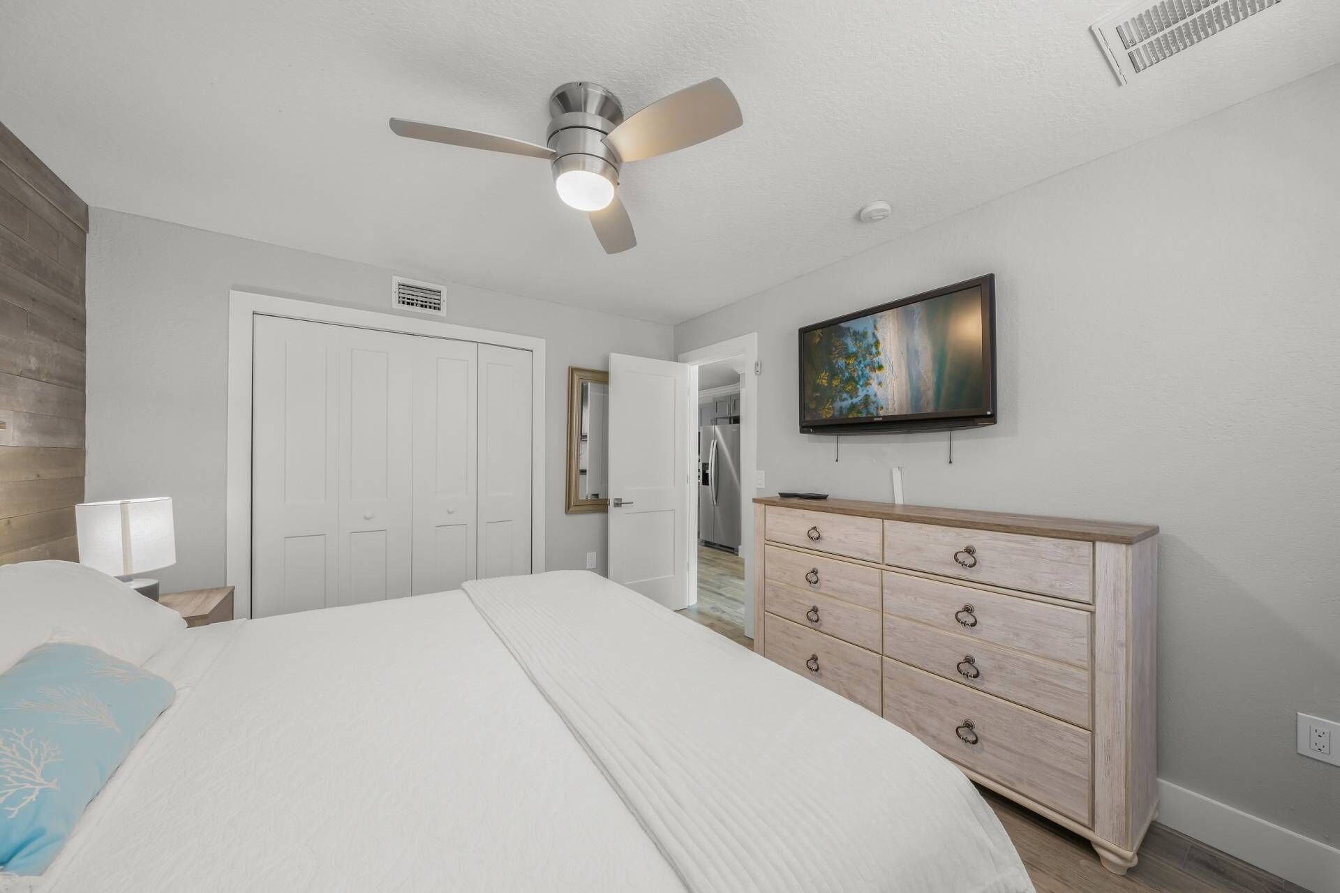 A bedroom with a bed, a dresser, a ceiling fan, a TV, and a door leading to a kitchen.