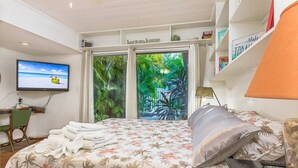 Een slaapkamer met een bed, een televisie, een bureau en uitzicht op een tropische tuin door een glazen deur.