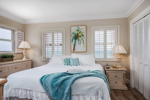 Quarto com cama grande, duas mesas de cabeceira, um criado-mudo, janela com persianas e um quadro de uma palmeira.
