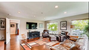A spacious living room with a leather sofa, a black entertainment unit, and a large window with a view of greenery. - Mid-Century 3 Bdr Lakewood Home Mountain Access! (Lakewood)