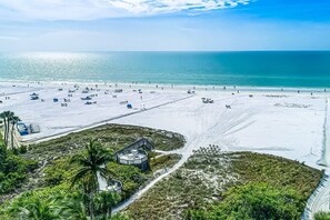 An aerial view of a beach with a large number of sunbeds and umbrellas, a small building with a circular deck, and a clear view of the ocean. - Serene Casarina (Siesta Key)