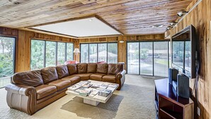 Amplia sala de estar con un sofá seccional de cuero marrón, una mesa de centro con revistas y un televisor sobre un mueble de madera. Grandes ventanas ofrecen luz natural y vistas a la vegetación exterior.