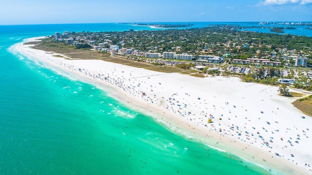 Vista aérea de un complejo turístico en la playa con una extensa franja de arena, aguas turquesas cristalinas y una zona residencial densa con edificios y vegetación al fondo.