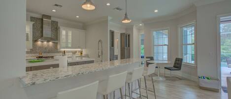 A modern kitchen with marble countertops, white bar stools, and pendant lighting.