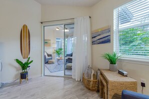 A modern living room with a glass door leading to a balcony, a surfboard on the wall, and a view of greenery through the window. - Siesta Bayside Condo 3 Bedroom (Siesta Key)