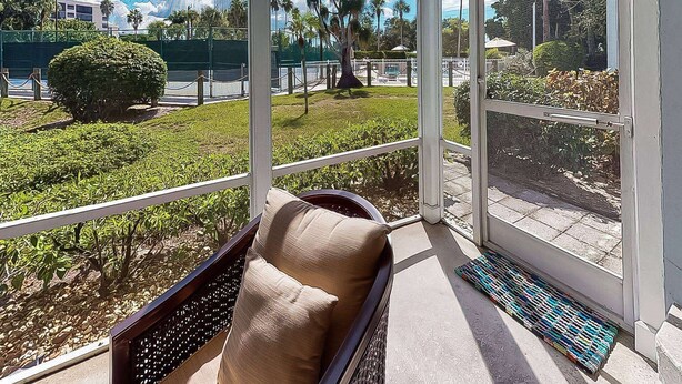 A balcony with a cushioned seat, a small table, and a view of a well-maintained garden with various plants and a palm tree. - Siesta Bayside by Pool & Tennis (Siesta Key)
