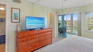 Un dormitorio con una cama grande, un aparador de madera con televisor de pantalla plana, un balcón con una silla y una puerta que da a otra habitación.