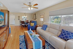A living room with a beige sofa, a coffee table, a dining area with chairs, a ceiling fan, and a window with blinds. - Siesta Harbor Portofino Canal View (Siesta Key)