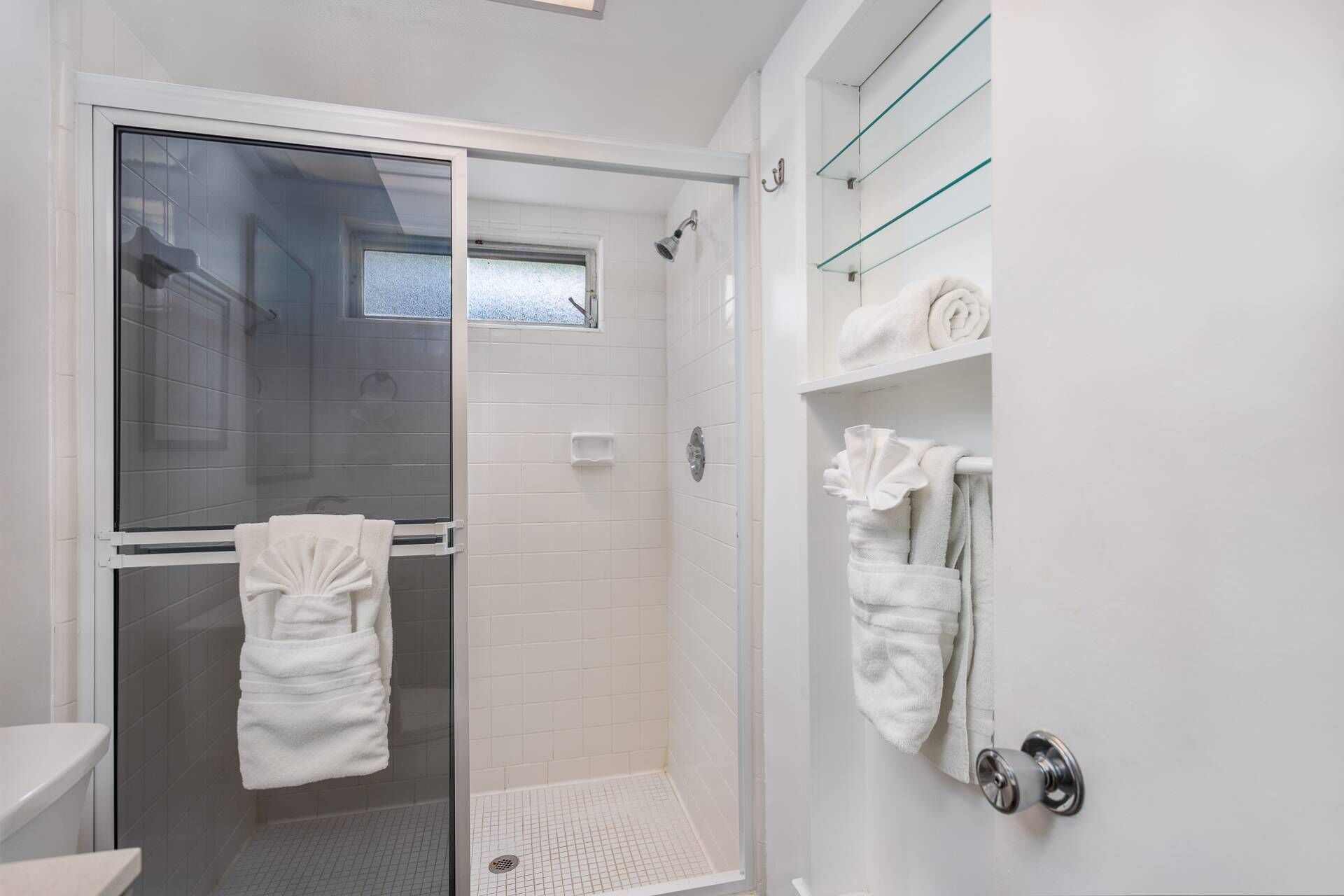A modern bathroom with a glass shower enclosure, white tiled walls, and a glass shelf with towels.