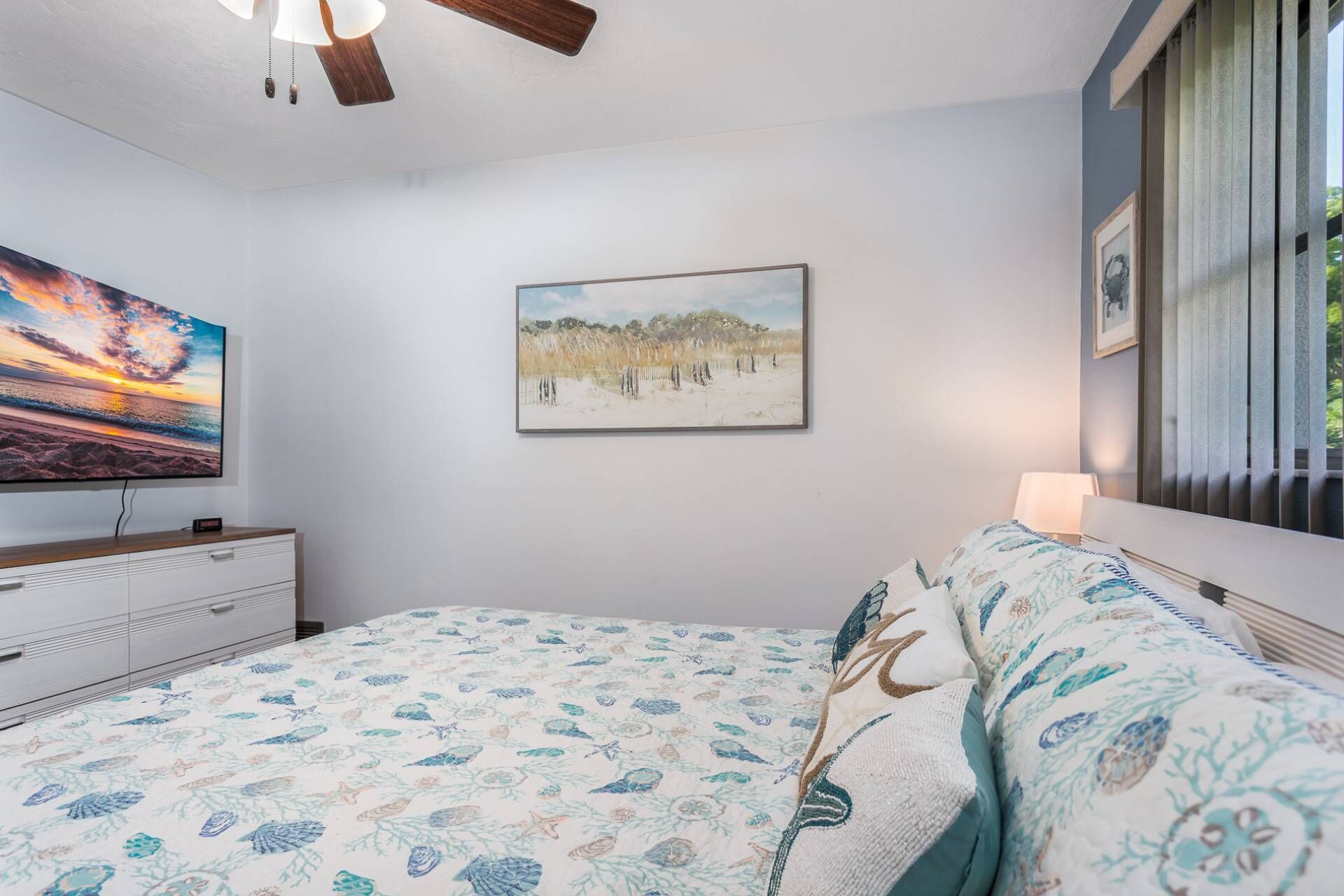 A bedroom with a bed, a painting of a beach, a TV on a dresser, and a window with blinds.