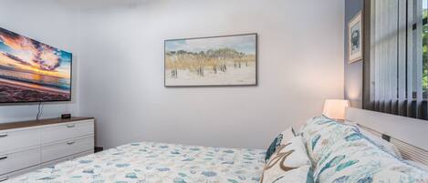 A bedroom with a bed, a painting of a beach, a TV on a dresser, and a window with blinds.