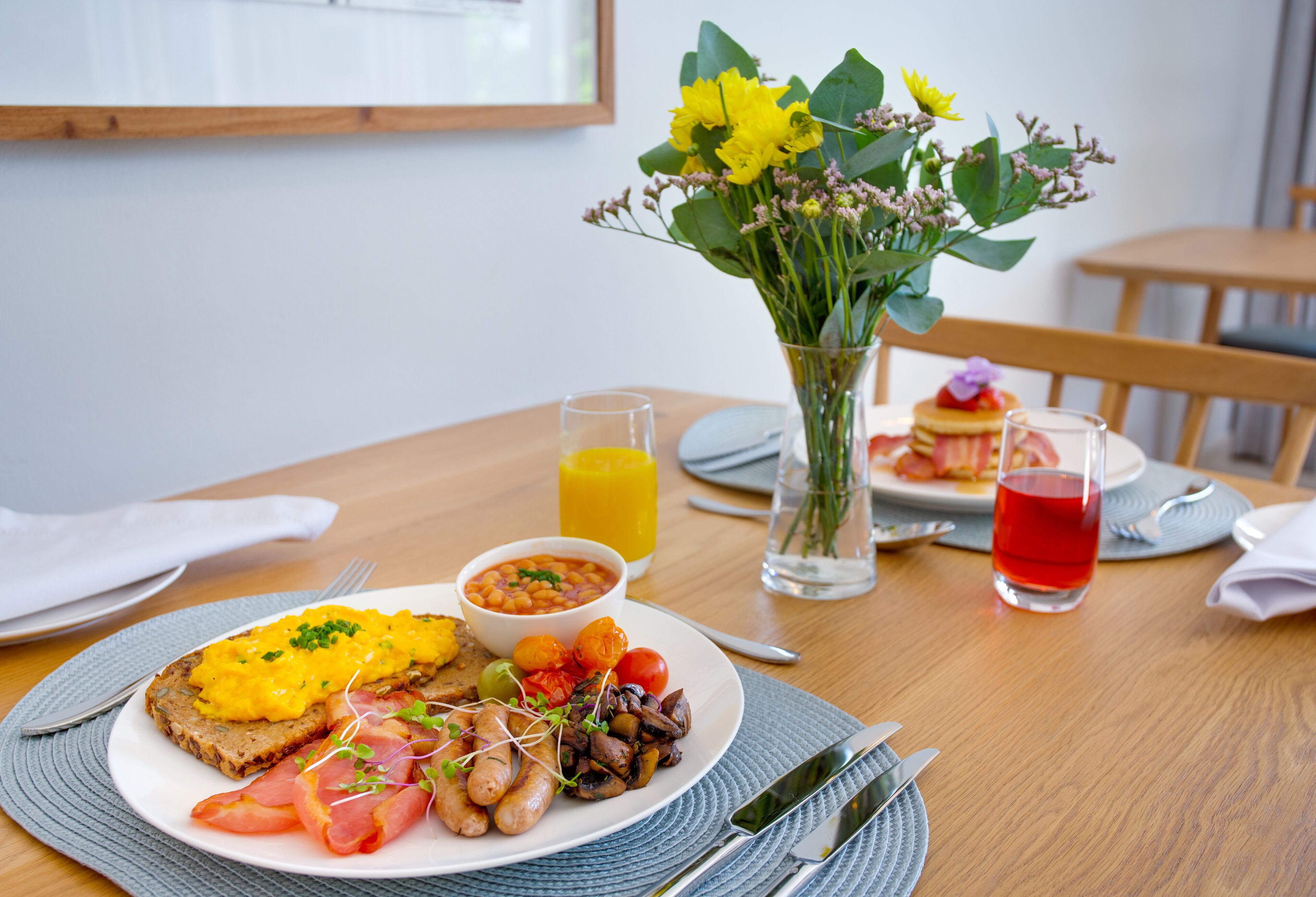 A breakfast setting with a plate of toast, eggs, tomatoes, and greens, a bowl of beans, a glass of orange juice, and a vase with flowers.