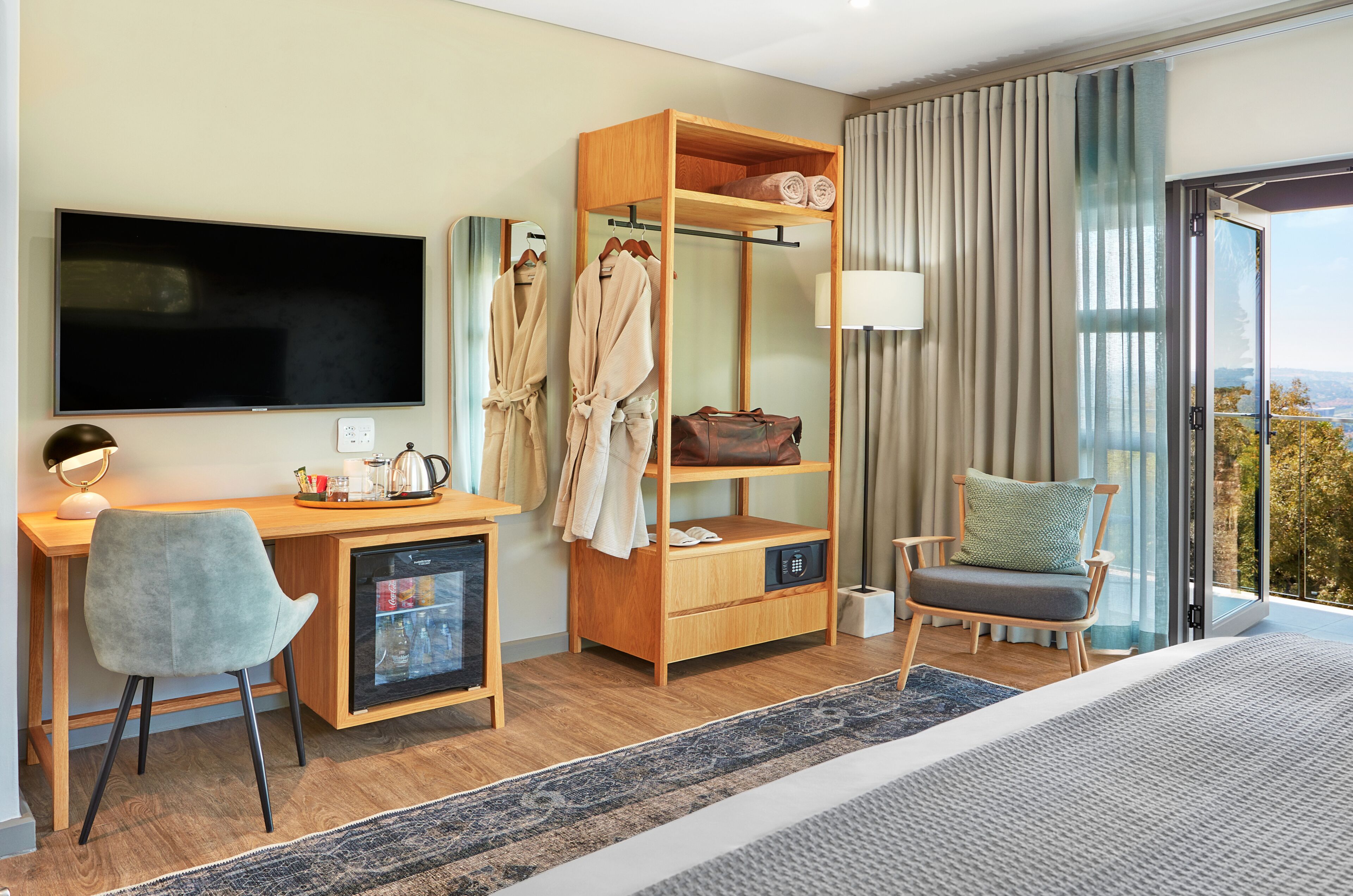 A modern hotel room with a large bed, a desk with a computer and a mini-fridge, a wardrobe, and a view of the outdoors.