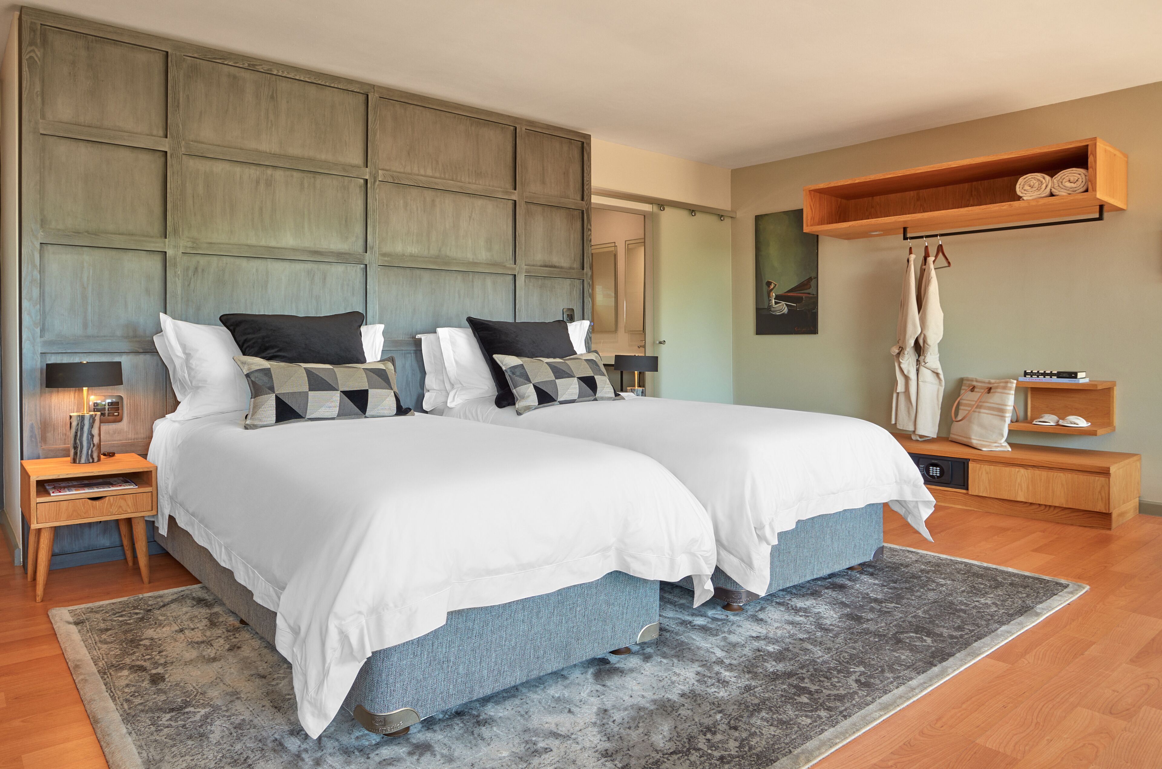 A modern bedroom with two beds, a wooden headboard, a bedside table with a lamp, a rug, and a wooden shelf with towels.