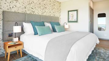 Una cama bien hecha con un cabecero gris, ropa de cama blanca y almohadas turquesa. Hay una mesita de noche con una lámpara y un libro. La habitación tiene papel tapiz con motivos florales y un cuadro en la pared.