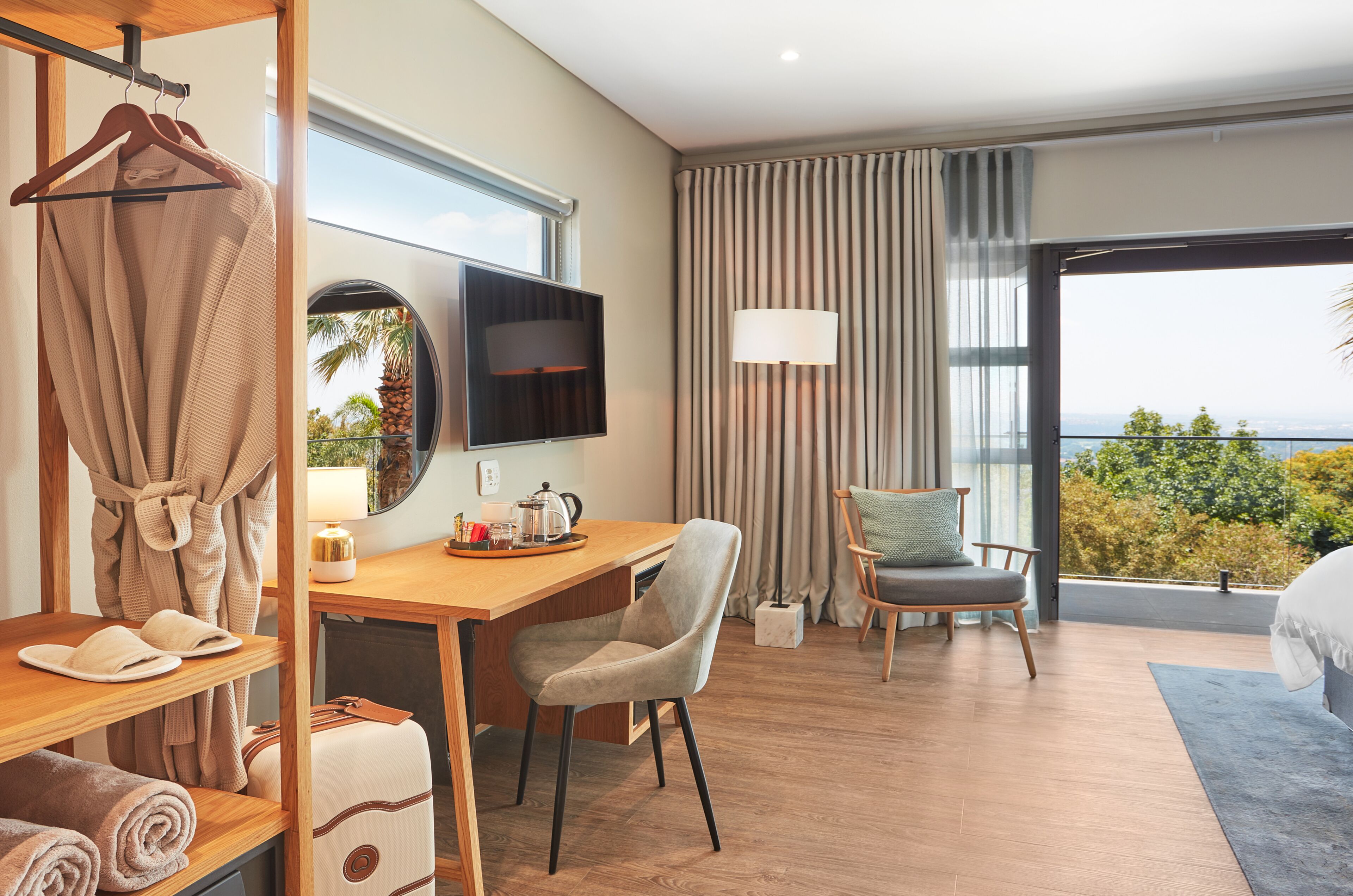 A hotel room with a wooden desk, a chair, a bed, a TV, and a view of the outdoors.