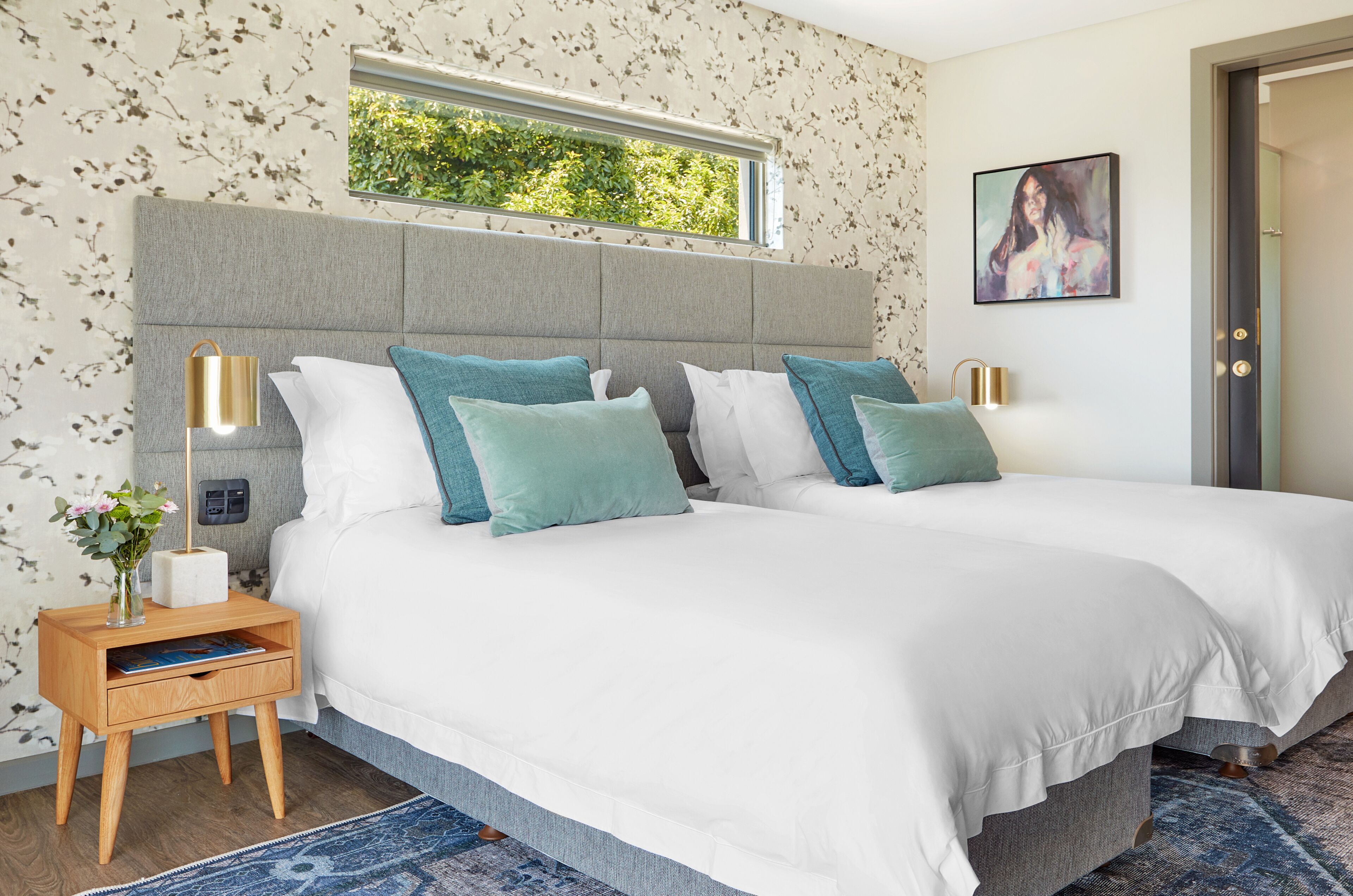 A double bed with white bedding and teal pillows, a bedside table with a lamp, and a window with a view of greenery.