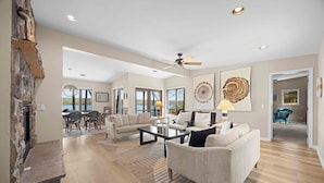 A spacious living room with a fireplace, a ceiling fan, and large windows offering a view of the outdoors.
