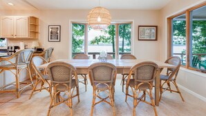 A dining area with a large table and wicker chairs, a hanging light fixture, and large windows with a view of greenery.