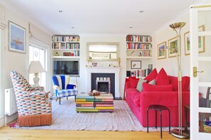 Ein Wohnzimmer mit Kamin, Bücherregalen, einem roten Sofa und einem gemusterten Sessel.