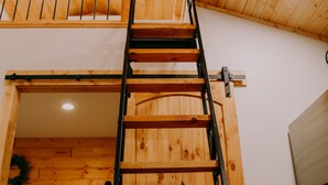 A wooden interior with a loft bedroom, a ladder, and a bed with a checkered blanket. - Newly Constructed! "The Stables" (Trenton)