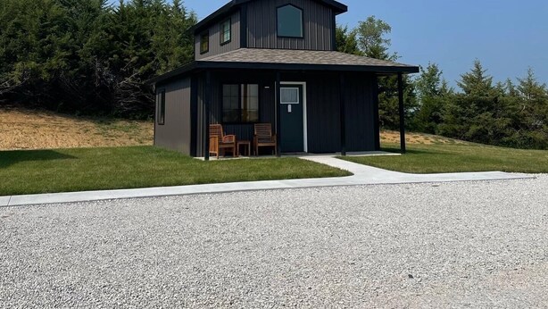 A modern two-story house with a dark exterior, a covered porch, and a gravel driveway. - Newly Constructed! "The Stables" (Trenton)