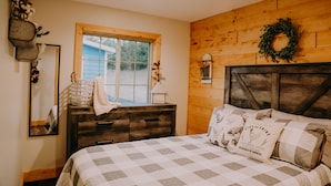 A bedroom with a wooden wall, a window, a dresser, a bed with a checkered bedspread, and a wall-mounted light fixture. - Newly Constructed! "The Stables" (Trenton)