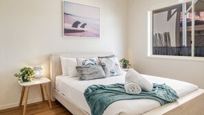 A bedroom with a bed, a bedside table with a plant, a wall-mounted artwork, and a window with a view of a building. - Beachside living takes a smart & stylish turn with this well appointed apartment (Lennox Head)