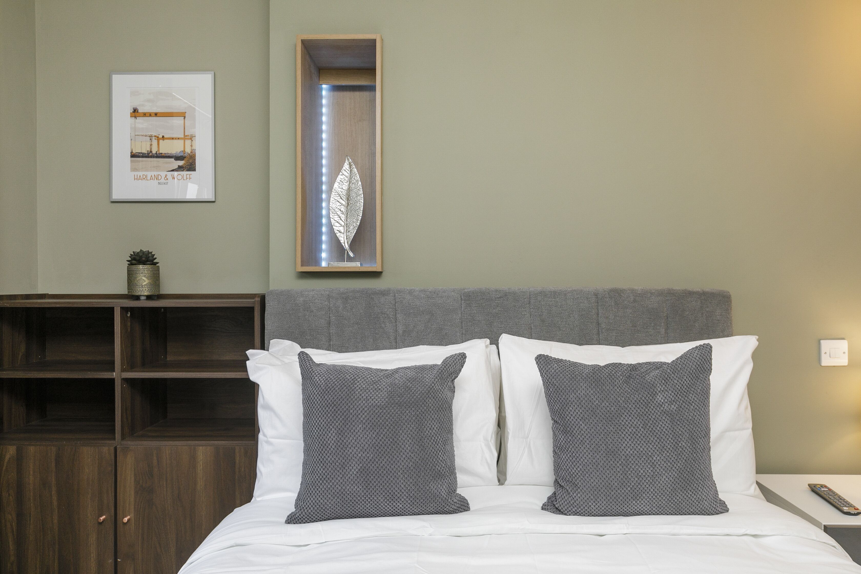 Una cama bien hecha con sábanas blancas y dos almohadas grises, un cuadro enmarcado encima y un mueble de madera debajo.