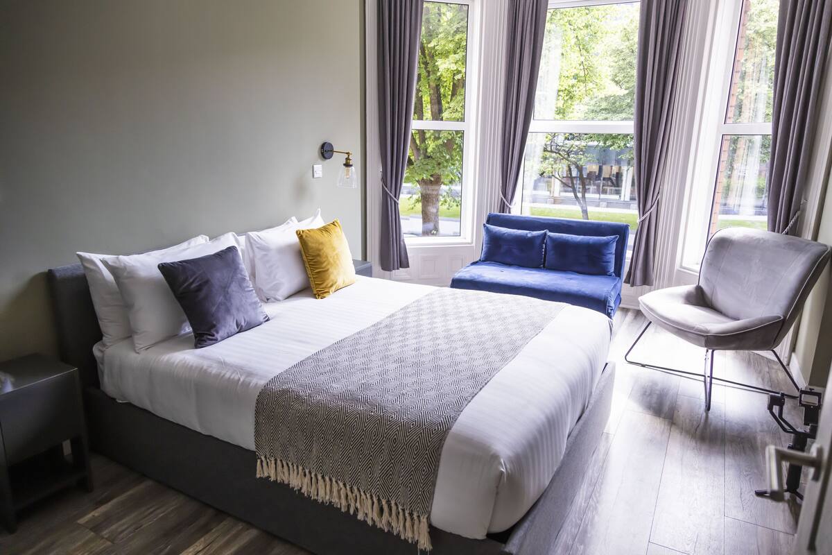 A modern bedroom with a large bed, a bedside table, a chair, and a view of the outdoors through large windows.