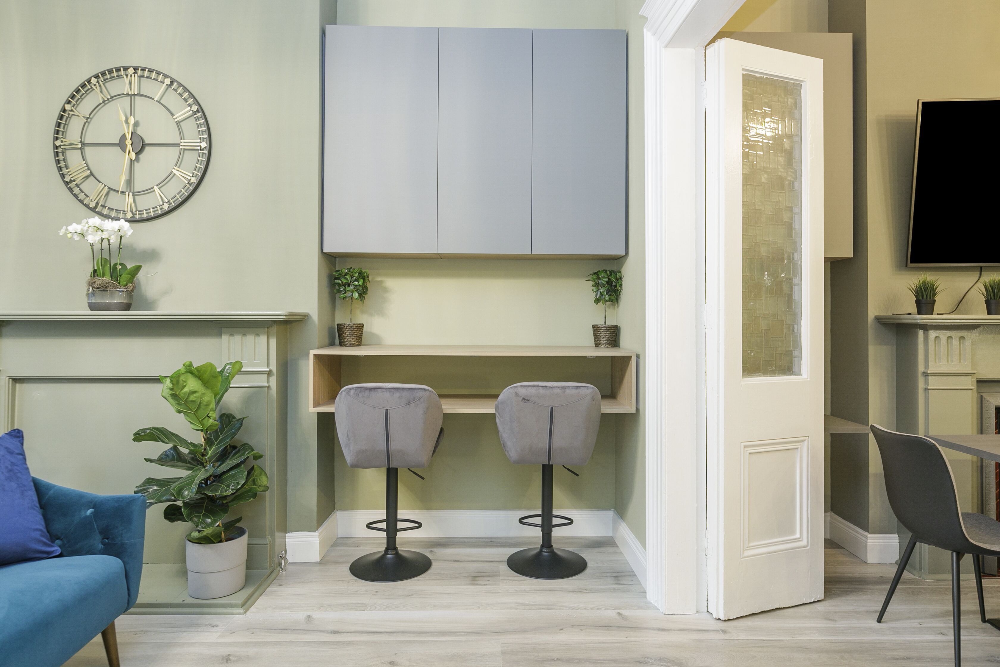 Un salon moderne avec un canapé bleu, deux tabourets de bar gris, une grande horloge murale et un téléviseur fixé au mur.